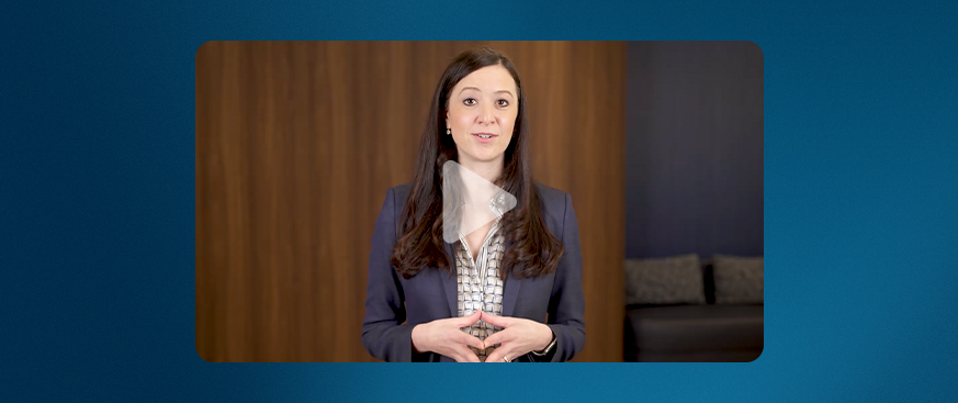 A woman with long dark hair, wearing a navy blazer over a patterned blouse, stands in front of a wooden wall and speaks while looking at the camera. A translucent play button overlay indicates this is a video still.
