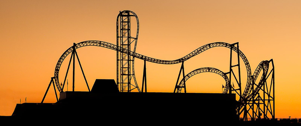 Silhouette of a roller coaster with loops and steep tracks, set against a bright orange sunset sky.