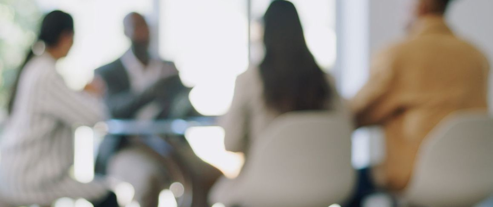 Four people sitting around a small round table in a modern office setting, engaged in a discussion. The image is intentionally blurred, with bright natural light coming through windows in the background.