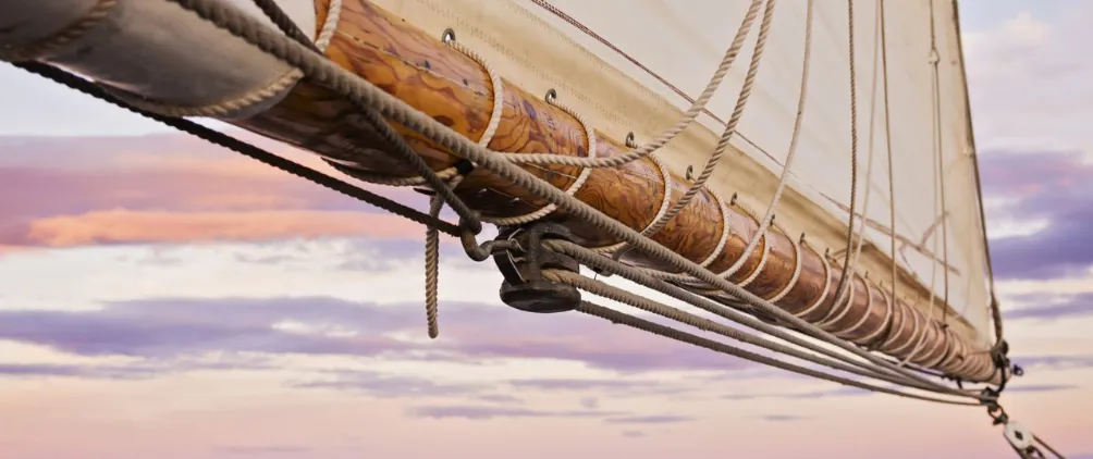 Close-up of a wooden boom with rigging and a sail attached, set against a pastel sky with soft pink and purple clouds at sunset.