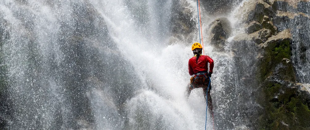Person wearing a helmet and climbing gear rappelling down a waterfall with water splashing around them.