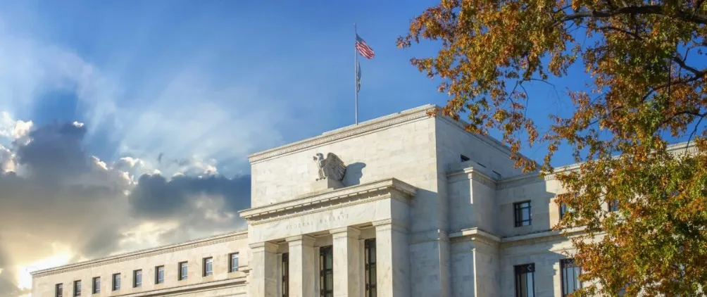 The Federal Reserve building in Washington, D.C., with the U.S. flag flying on top, sunlight streaming through clouds in the background, and autumn tree branches on the right.