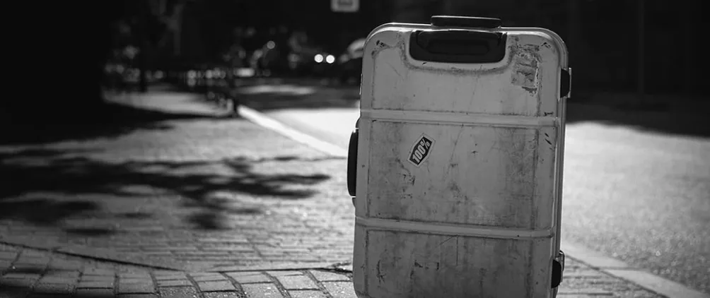 Worn hard-shell suitcase standing upright on a sunlit city sidewalk, black-and-white street scene suggesting travel, loss, or urban journey.