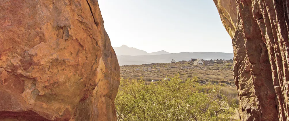 Desert landscape framed by two large reddish rock formations, overlooking a dry valley with scattered shrubs, distant mountains, and a small rural settlement under a clear sky